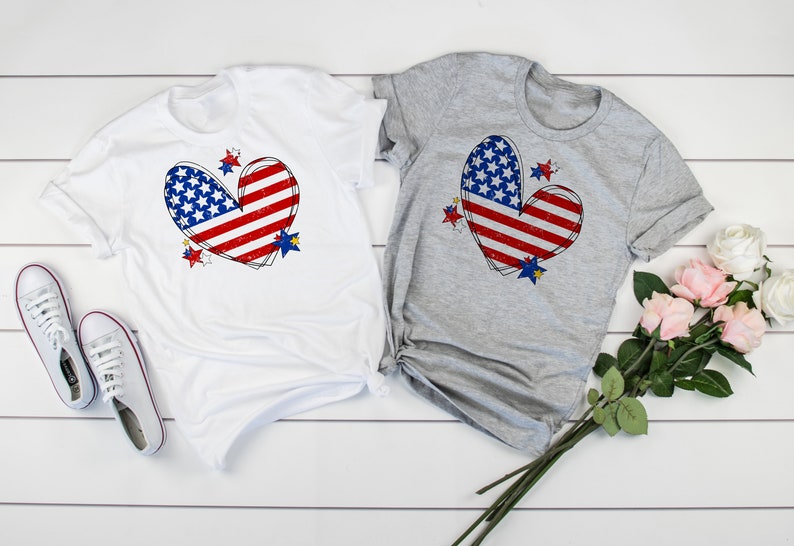 May include: Two t-shirts with a red, white, and blue heart-shaped American flag design. The shirts are white and grey and have a distressed look.