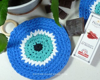 May include: Blue crocheted trivet with a white and turquoise eye design. The trivet is sitting on a white surface next to a package of chocolate with the text "Cioccolato di Modica al Peperoncino" and a red chili pepper graphic.