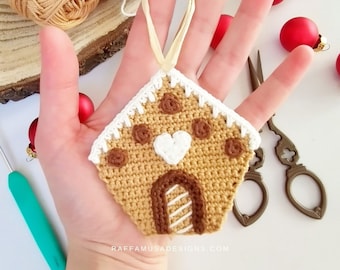 Puede incluir: Un adorno de casa de jengibre tejido a crochet con detalles de glaseado marrón, un corazón blanco y una puerta a rayas. El adorno está colgado de una cuerda y se sostiene en una mano.
