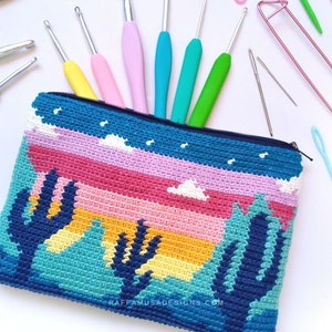 Puede incluir: Una bolsa de cremallera de ganchillo con un diseño de puesta de sol y cactus coloridos. La bolsa está hecha con una variedad de colores, incluyendo azul, verde, amarillo, naranja, rosa y rojo. El texto "RAFFAMUSADESIGNS.COM" está impreso en la parte inferior de la bolsa.