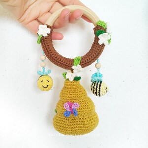 May include: A crocheted beehive toy with a wooden ring and hanging bee and flower charms. The beehive is yellow and has a pink butterfly on it. The ring is brown and has green leaves and white flowers around it.