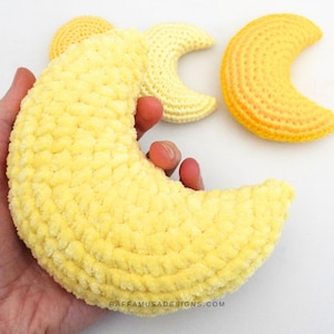 May include: Three crocheted crescent moon shapes in yellow and orange shades. The largest moon is made with a fuzzy yarn and is being held in a hand.