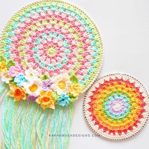 May include: Two crocheted dreamcatchers with rainbow colors and floral accents. The larger dreamcatcher has a fringe of yarn and is decorated with crocheted flowers in shades of pink, yellow, and blue. The smaller dreamcatcher is a solid circle with a rainbow pattern.