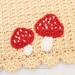 Può includere: Primo piano di un pezzo all'uncinetto con due funghi rossi con pois bianchi e gambi. I funghi sono su uno sfondo color crema con un bordo smerlato. Il testo "RAFFAMUSADESIGNS.COM" è visibile.