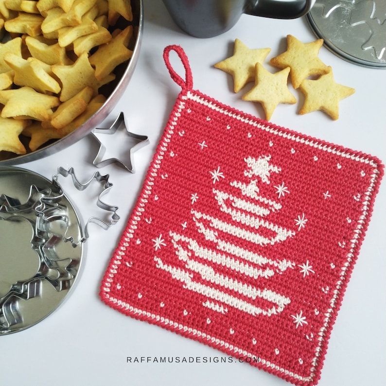 K&ouml;nnte beinhalten: Ein roter geh&auml;kelter Topflappen mit einem wei&szlig;en geh&auml;kelten Weihnachtsbaum-Design. Der Topflappen hat eine Schlaufe zum Aufh&auml;ngen. Der Text "RAFFAMUSADESIGNS.COM" befindet sich am unteren Rand des Topflappens.
