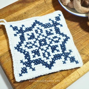 May include: White and navy blue knitted potholder with a geometric design. The potholder has a loop for hanging and is sitting on a wooden cutting board. The text "RAFFAMUSADESIGNS.COM" is visible on the bottom edge.