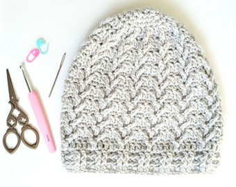 May include: A gray crocheted beanie with a textured stitch pattern. The beanie is on a white surface with a pair of scissors, a crochet hook, and stitch markers.