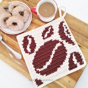 Puede incluir: Un salvamanteles cuadrado de ganchillo color crema con un diseño de granos de café marrones. El salvamanteles está sobre una tabla de cortar de madera, junto a un plato de bagels y una taza de café. El salvamanteles tiene un lazo para colgar.