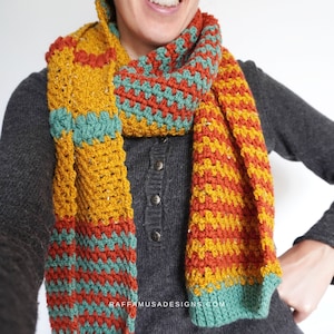 May include: A crocheted scarf with stripes of mustard yellow, burnt orange, and teal green. The scarf is folded in half and draped around a person's neck.