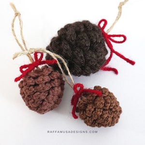 May include: Three handmade crochet pinecone ornaments in varying shades of brown. Each pinecone is adorned with a small red bow and a natural twine loop for hanging. The ornaments are displayed on a white background.