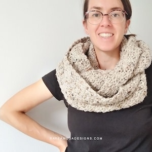 May include: A woman wearing a beige crocheted infinity scarf with black flecks. The scarf is worn around the neck and drapes down the front of the chest. The woman is smiling and wearing glasses.