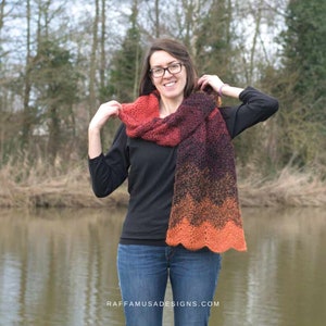 May include: A hand-knitted scarf in shades of red, purple, and orange. The scarf has a wavy, scalloped edge and is wrapped around the neck. The scarf is made of a soft, textured yarn. The website address raffamusadesigns.com is visible.