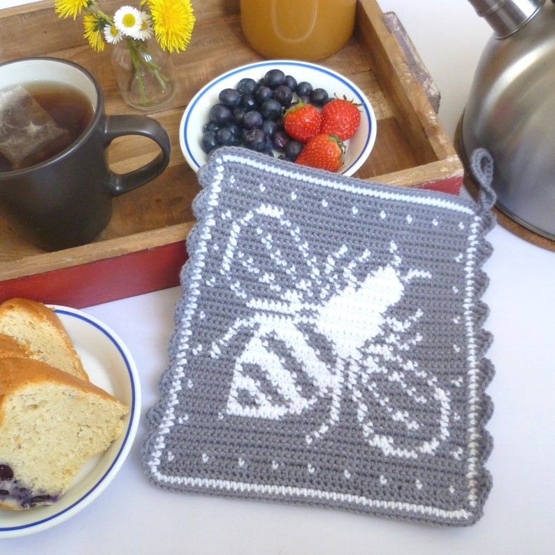 K&ouml;nnte beinhalten: Grauer geh&auml;kelter Topflappen mit einem wei&szlig;en Bienen-Design. Der Topflappen hat einen gewellten Rand und eine Schlaufe zum Aufh&auml;ngen.