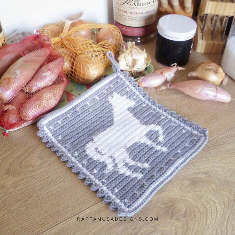 K&ouml;nnte beinhalten: Grauer H&auml;kel-Topflappen mit einem wei&szlig;en Einhorn-Design. Der Topflappen hat einen gewellten Rand und eine Schlaufe zum Aufh&auml;ngen.