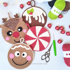 Puede incluir: Cinco posavasos de Navidad hechos a crochet, incluyendo un hombre de jengibre, un reno, un bastón de caramelo y un pudín de Navidad. Los posavasos están sobre una superficie blanca con adornos rojos y verdes.