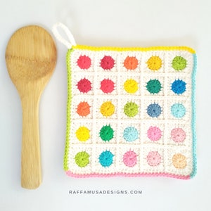 May include: A colorful crochet pot holder with a white background and a pink, yellow, and green border. The pot holder has a loop for hanging.