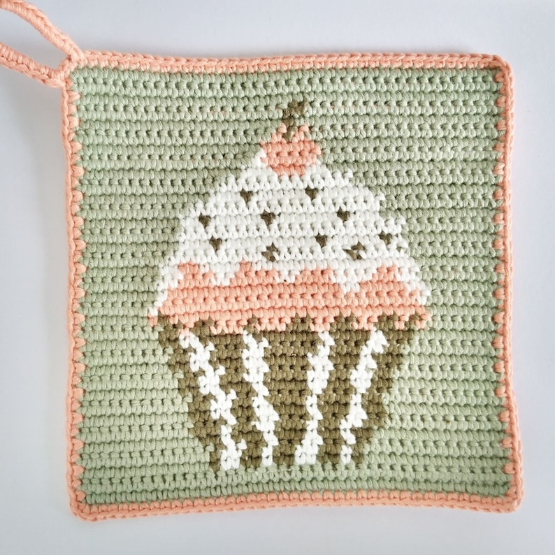 May include: A green crocheted pot holder with a pink and white cupcake design. The cupcake has brown frosting and white sprinkles.