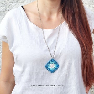 Può includere: Una collana con catena d'argento con un ciondolo quadrato a uncinetto blu, bianco e azzurro. Il ciondolo ha una forma di diamante. La collana è indossata da una persona con i capelli lunghi castani. Il testo "RAFFAMUSADESIGNS.COM" è visibile nella parte inferiore dell'immagine.