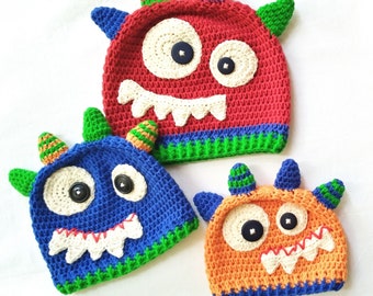 Könnte beinhalten: Drei gehäkelte Monstermützen in Rot, Blau und Orange mit weißen Augen und Zähnen. Die Mützen haben grüne und gelbe Akzente.