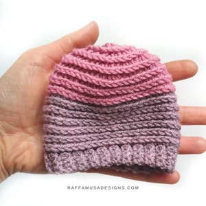 May include: A pink and purple knitted hat with a ribbed texture. The hat is held in a hand.