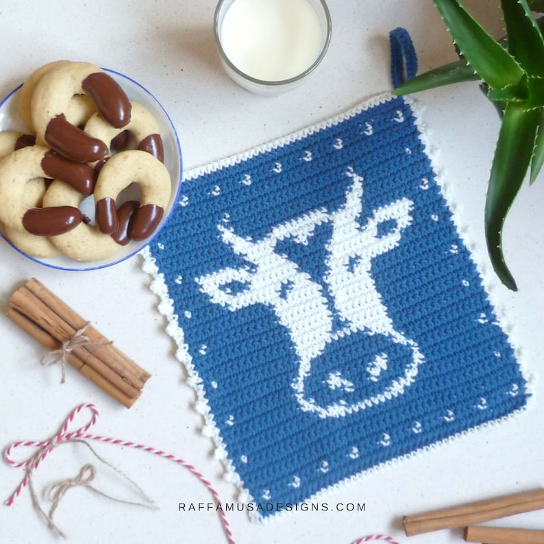 K&ouml;nnte beinhalten: Ein blauer H&auml;kel-Topflappen mit einem wei&szlig;en Kuh-Design. Der Topflappen liegt auf einer wei&szlig;en Oberfl&auml;che mit Zimtstangen, Schnur und einer Sch&uuml;ssel mit Keksen.
