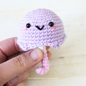 May include: A crocheted purple jellyfish with a pink tail and black eyes. The jellyfish has a happy face with a small black smile.