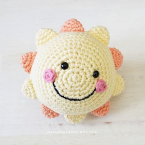 Pode incluir: Um brinquedo de sol de crochê com um corpo amarelo, raios cor de pêssego e um rosto sorridente com olhos pretos e bochechas cor-de-rosa.