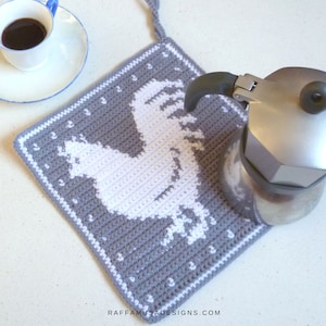 K&ouml;nnte beinhalten: Grauer geh&auml;kelter Topflappen mit einem wei&szlig;en Hahn-Design. Der Topflappen hat eine Schlaufe zum Aufh&auml;ngen. Eine silberne Kaffeekanne steht auf dem Topflappen.