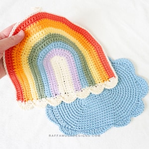 May include: A crocheted rainbow with a white cloud border and a blue crocheted cloud. The rainbow is made with different colored yarn, including red, orange, yellow, green, blue, and purple.