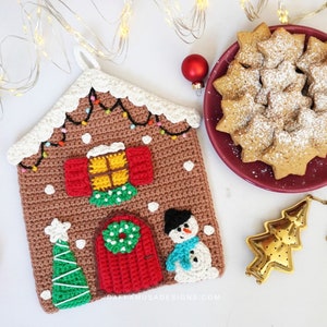 Puede incluir: Manopla de cocina de jengibre tejida a crochet con un muñeco de nieve, un árbol de Navidad y una corona en la puerta. La manopla es marrón con glaseado blanco y luces de colores. El fondo incluye un plato de galletas con forma de estrella y un infusor de té dorado.