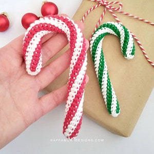 Puede incluir: Dos bastones de caramelo tejidos a crochet, uno rojo y blanco y otro verde y blanco, se sostienen en una mano. Los bastones de caramelo están sobre un fondo de papel marrón.