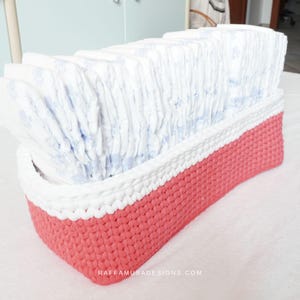 Puede incluir: Una cesta de crochet blanca y coral llena de una pila de pañales de bebé blancos. La cesta está hecha con un punto de crochet texturizado y tiene una banda superior blanca y una banda inferior coral. El texto "RAFFAMUSADESIGNS.COM" es visible en la parte inferior de la imagen.