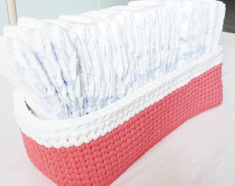 以下が含まれることがあります： 白と珊瑚色の編み物のかごに、白いベビーおむつが積み重ねられています。かごは、質感のある編み目の編み方でできており、白い上部バンドと珊瑚色の下部バンドが付いています。画像の下部に「RAFFAMUSADESIGNS.COM」というテキストが表示されています。