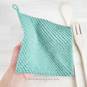 May include: A light green knitted dishcloth with a loop handle. The dishcloth is made with a textured stitch pattern. The dishcloth is lying on a white wooden surface.