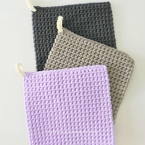 Pode incluir: Três panos de crochê feitos à mão em tons de cinza, bege e lavanda. Cada pano tem uma alça para pendurar. O ponto de crochê cria uma superfície texturizada. Os panos são exibidos em um fundo branco.