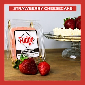 May include: A clear plastic container of strawberry cheesecake fudge with a label reading Fudge etc. Strawberry Cheesecake 8 oz. (226.8g). Two fresh red strawberries sit in front of the container, and a cheesecake on a glass stand is visible.