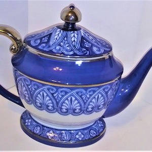 May include: A blue and white ceramic teapot with a gold handle and accents. The teapot features a decorative floral pattern and a gold-colored lid. The spout and handle are a deep blue, and the base is a matching design.