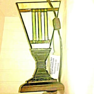 May include: Mission Tiffany style upright accent lamp with a stained glass shade. The shade features white, green, and brown panels. The lamp has a dark bronze base and a power cord with a plug. The tag reads: Mission Tiffany Style Upright Accent Lamp Requires one 40-watt bulb.