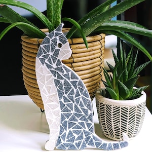 May include: A mosaic cat figurine in black and white, sitting on a white surface. The cat is surrounded by two potted plants with green leaves.