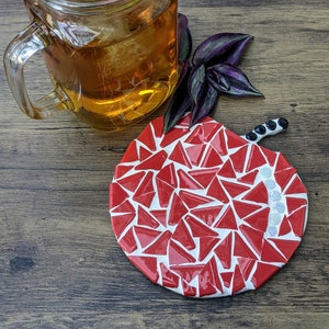 May include: A red and white mosaic apple-shaped coaster with a black stem. The coaster is made of small, triangular pieces of tile.