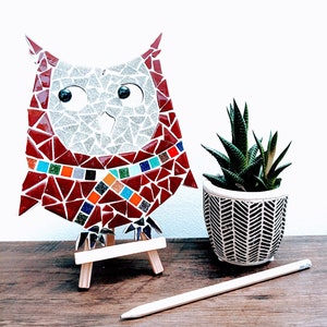 Può includere: Una statuetta di gufo in mosaico rosso, bianco e blu su un cavalletto di legno. Il gufo ha grandi occhi rotondi e un piccolo becco. Il gufo è circondato da una pianta grassa verde in un vaso con motivo bianco e nero.