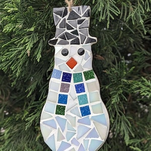 Könnte beinhalten: Ein Mosaik-Schneemann-Anhänger mit einem schwarzen Zylinder, einer roten Nase und einem blau-grünen Schal. Der Schneemann besteht aus weißen, blauen, grünen und schwarzen Mosaikfliesen.