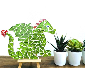 May include: A mosaic dragon figure crafted from triangular green glass pieces with red accents on the tail and spine, featuring a single blue eye. The dragon is mounted on a small wooden easel stand, positioned next to two small potted succulents.