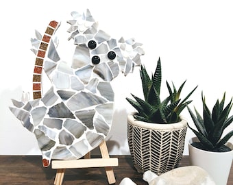 May include: A gray mosaic koala sculpture on a wooden easel. The koala is made of small, irregular pieces of glass in shades of gray and white. The koala has black eyes and a red tail. The sculpture is surrounded by white stones.