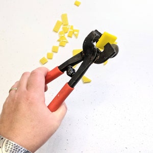 May include: A hand holding a pair of black and red glass tile nippers with a yellow tile in the jaws. The nippers are used to cut small pieces of glass tile for mosaic projects.