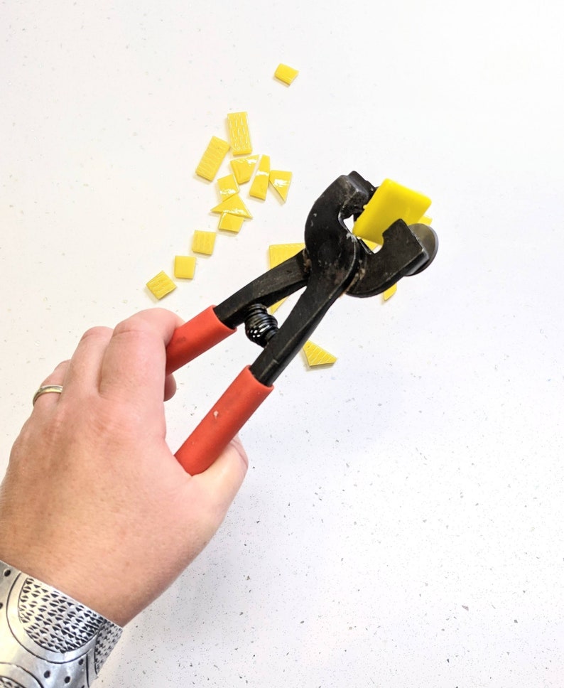 May include: A hand holding a pair of black and red glass tile nippers with a yellow tile in the jaws. The nippers are used to cut small pieces of glass tile for mosaic projects.