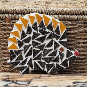 May include: A mosaic hedgehog figure crafted from white grout and triangular glass tiles. The body features dark gray textured pieces, while the upper back is lined with bright orange triangles. A brown circular tile serves as the eye and a red tile as the nose.