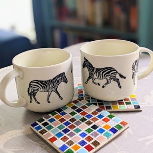 May include: Two white ceramic mugs with black zebra illustrations on a colourful mosaic tile coaster set.