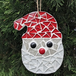 May include: A white and red mosaic Christmas ornament shaped like Santa Claus's face. The ornament has a red hat, white beard, and black eyes.
