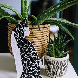 Könnte beinhalten: Eine Mosaik-Katzenfigur in Schwarz und Weiß mit einem hellblauen Auge steht auf einer weißen Oberfläche. Dahinter befinden sich zwei Topfpflanzen: eine große Aloe Vera in einem geflochtenen Korb und eine kleine Sukkulente in einem gemusterten Topf. Dekorativer Artikel.